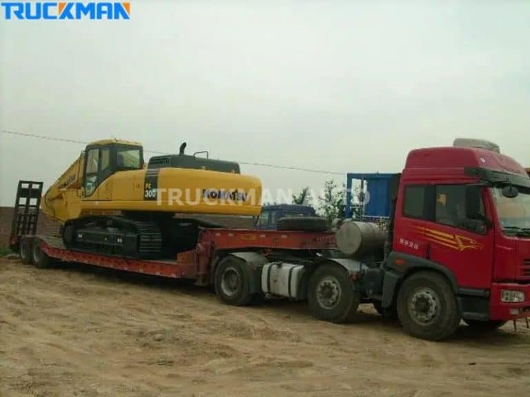 Pesada excavadora amarilla transportada en un semirremolque rojo de plataforma baja por un camino de tierra