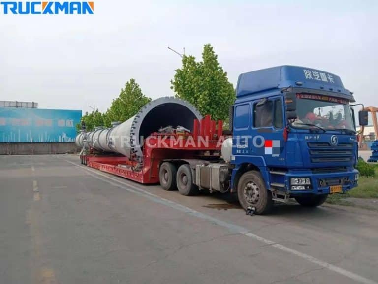 Camión azul de gran tonelaje con semirremolque de plataforma baja que transporta equipos industriales cilíndricos de gran tamaño por las carreteras de la ciudad.