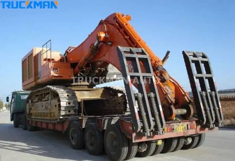 Semirremolque de plataforma baja con rampas desplegadas cargando una excavadora de orugas naranja.