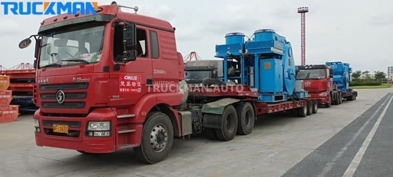 Semirremolque rojo de plataforma baja transportando grandes equipos industriales azules en una terminal portuaria